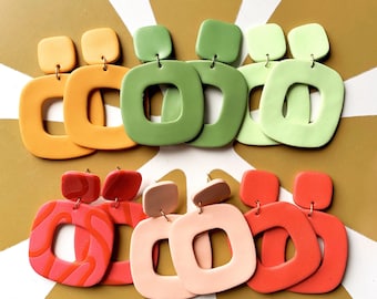 May include: A collection of colorful, square-shaped earrings in various shades of orange, green, pink, and red. Each pair features a smaller square top and a larger, open-center square design. The earrings are displayed on a gold and white striped surface.