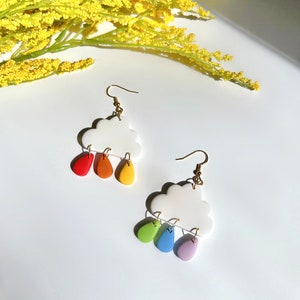 May include: A pair of white cloud-shaped earrings with rainbow-colored raindrop charms. The earrings are made of polymer clay and have gold hooks.