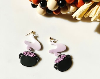 May include: A pair of whimsical earrings featuring a black cauldron with purple bubbles and a light purple, cloud-like shape. The earrings have gold-colored posts and are set against a white background. Decorative wooden beads and dried flowers are in the background.
