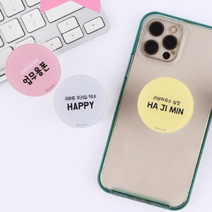 May include: Four colourful phone grips with different text and designs. The grips are pink, white, yellow, and blue. The text on the grips reads "업무용폰", "HAPPY", "HA JI MIN", "KIM JI MIN 010,2345,6789", and "김DANA".