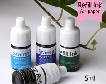 May include: Three small bottles of ink for paper, labeled "Stamp Refill Ink" in blue, violet, and green. The bottles are 5 ml each and are on a white surface.