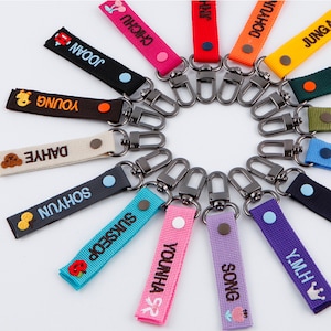 May include: A set of 12 colorful keychains with embroidered names and small decorative details. Each keychain has a silver metal hook.