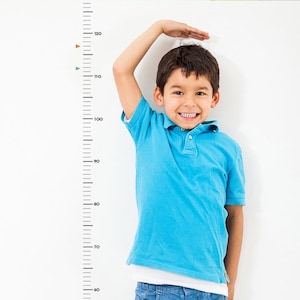 May include: A young boy stands in front of a white wall with a black and white height chart. He is wearing a blue polo shirt and blue jeans. The chart shows measurements in centimetres from 60 to 120.