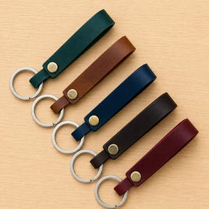 May include: Five leather keychains arranged diagonally, each featuring a folded strap in dark green, tan, navy blue, dark brown, or burgundy. Each keychain is secured with a brass-toned metal rivet and includes a circular silver-toned metal split ring.
