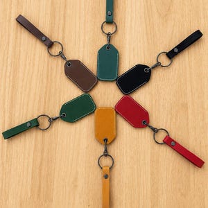 May include: Six leather keychains arranged in a star pattern. Each keychain features a rectangular tag with rounded corners, white stitching, a metal split ring, and a matching leather strap secured with a dark metal snap button fastener.