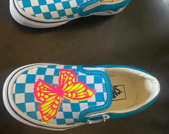 May include: A pair of blue and white checkered Vans slip-on shoes with a bright pink and yellow butterfly design on one shoe. The shoes have a white sole and a blue inner lining.