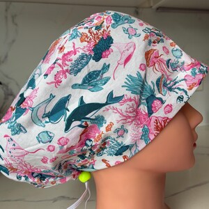 May include: White scrub cap with a vibrant ocean design. The cap is patterned with illustrations of dolphins, sea turtles, jellyfish, and coral in teal, pink, and orange. It is secured with a yellow toggle and white elastic.