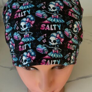 May include: Black scrub cap with a repeating pattern of skeleton figures holding salt shakers. The figures wear blue tutus and pink bows. Text includes "DON'T BE SALTY" and "SALTY" in pink and blue.