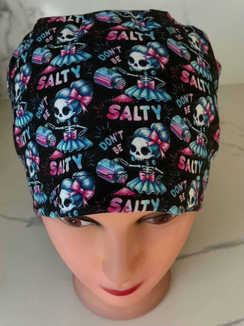 May include: Black scrub cap with a repeating pattern of skeleton figures holding salt shakers. The figures wear blue tutus and pink bows. Text includes "DON'T BE SALTY" and "SALTY" in pink and blue.