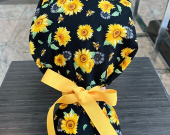 May include: A black scrub hat with a sunflower and bee print. The hat is tied with a yellow ribbon.