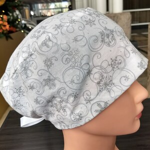 May include: A white scrub hat with a silver snowflake and swirl pattern. The hat has a white ribbon tie.