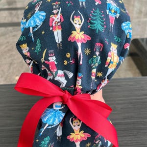 May include: A black scrub hat with a red ribbon tied around it. The hat features a repeating pattern of ballerinas and nutcracker soldiers on a dark background.