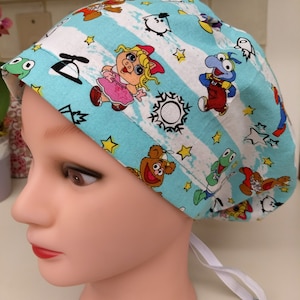 May include: A turquoise blue scrub hat with a white stripe pattern. The hat features a variety of cartoon characters, including Miss Piggy, Cookie Monster, and Kermit the Frog.