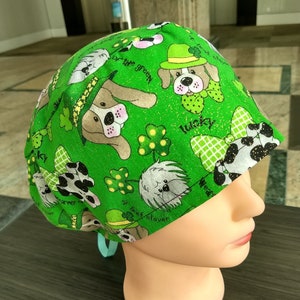May include: Green surgical scrub hat with a fun St. Patrick's Day theme. The hat features a green background with a variety of dogs wearing shamrock hats and the text "Wearin' of the Green", "Lucky", and "Four Leaf Clover".
