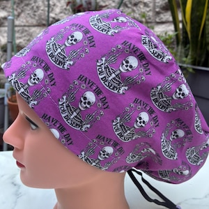 May include: A purple surgical scrub cap with a repeating pattern of white skeletons and the text "HAVE THE DAY YOU DESERVE". The cap has a black tie closure and is worn on a mannequin head.