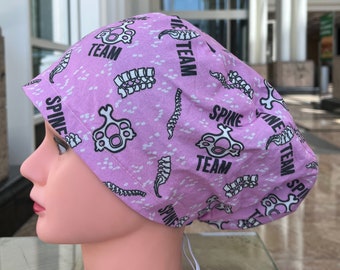 May include: Pink surgical scrub hat with a white and black print of vertebrae and the words "Spine Team" repeated throughout.