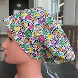 Könnte beinhalten: Eine weiße OP-Haube mit einem bunten Muster aus Regenbogen, Sternen, Herzen, Kleeblättern und dem Buchstaben "S". Die Haube hat ein Gummiband für einen sicheren Sitz. Das Design weist Pastellfarben auf.
