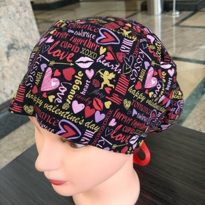 May include: A black surgical cap with a Valentine's Day theme. The cap is covered in hearts, lips, and the words "love," "kisses," and "happy Valentine's Day" in various colors. A red ribbon is attached to the back.
