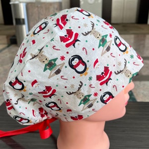Può includere: Un cappello da lavoro bianco con una stampa a tema natalizio con pinguini, renne, Babbo Natale, bastoncini di zucchero e fiocchi di neve. Il cappello ha lacci rossi.