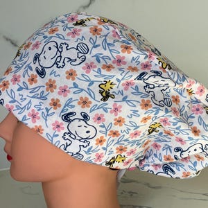 May include: A white scrub cap featuring a repeating pattern of Snoopy, Woodstock, and floral elements in pink, orange, and blue. The cap is designed with a rounded crown and a comfortable fit, suitable for medical professionals or anyone seeking a stylish head covering.