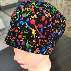May include: A black scrub hat with a colourful paint splatter pattern. The hat is made of fabric and has a tie at the back.