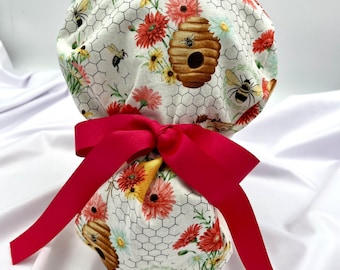 May include: A white scrub hat with a beehive, flowers, and honeycomb pattern. The hat is tied with a bright pink ribbon.