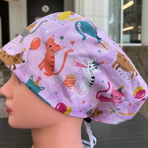 May include: A purple scrub hat with a colorful pattern of cats and dogs wearing party hats and holding balloons and presents. The hat has a white drawstring.