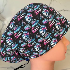 May include: A black surgical scrub cap with a repeating pattern of skeleton figures with pink bows and blue skirts, holding a pink and blue water bottle. The words "DON'T BE SALTY" and "SALTY" are also printed on the cap.