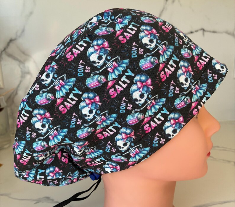 May include: A black surgical scrub cap with a repeating pattern of skeleton figures with pink bows and blue skirts, holding a pink and blue water bottle. The words "DON'T BE SALTY" and "SALTY" are also printed on the cap.