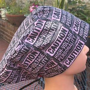 Può includere: Un cappello chirurgico nero e rosa con la scritta ripetuta "CAUTION HIGH-VOLTAGE PERSONALITY". Il cappello è fatto di tessuto ed è progettato per coprire la testa. Il cappello è indossato da un manichino.