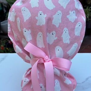 May include: A pink scrub cap featuring a pattern of white cartoon ghosts. The cap is tied with a pink ribbon. The fabric is a light pink color, and the ghosts have simple, smiling faces.