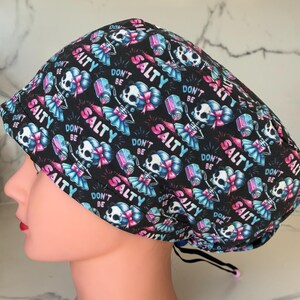 May include: Black surgical scrub cap with a repeating pattern of skeleton figures, salt shakers, and the words "DON'T BE SALTY" in pink and teal. The cap has a black drawstring for adjustment.