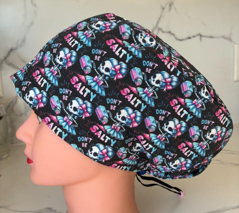 May include: Black surgical scrub cap with a repeating pattern of skeleton figures, salt shakers, and the words "DON'T BE SALTY" in pink and teal. The cap has a black drawstring for adjustment.