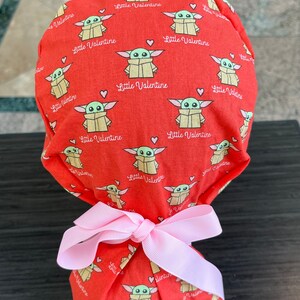 Peut inclure: Un sac cadeau en tissu rouge avec un ruban rose. Le sac est décoré d'un motif répété de personnages de bébé Yoda verts avec le texte "Little Valentine" sur un fond rouge.