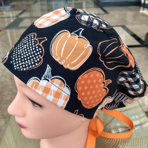 Peut inclure: Un bonnet de travail noir avec un imprimé de citrouille en orange, blanc et noir. Le bonnet a une cravate orange.