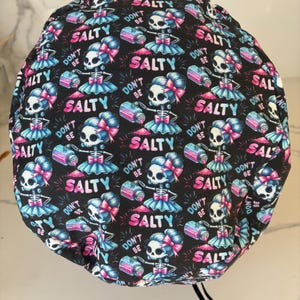 May include: Black surgical scrub cap with a repeating pattern of cartoon skeletons in tutus, holding salt shakers. The design includes the phrases "DON'T BE SALTY" and "SALTY" in pink and blue.