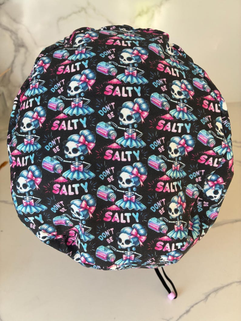May include: Black surgical scrub cap with a repeating pattern of cartoon skeletons in tutus, holding salt shakers. The design includes the phrases "DON'T BE SALTY" and "SALTY" in pink and blue.