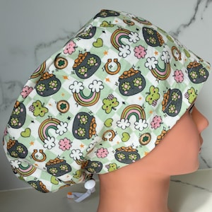 Könnte beinhalten: Eine gemusterte OP-Haube mit St. Patrick's Day-Motiv. Die Haube hat einen weiß-grünen Karohintergrund mit Illustrationen von Regenbogen, Goldtöpfen, Kleeblättern und Hufeisen. Die Haube ist so konzipiert, dass sie bequem und sicher sitzt.