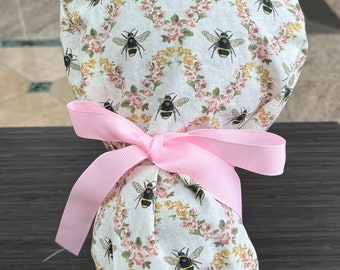 May include: A white scrub hat with a pink ribbon tied around it. The hat has a floral pattern with bees on it.