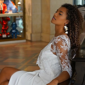 Puede incluir: Una mujer con un top blanco de encaje y una falda blanca se sienta en un banco de piano. Lleva tacones blancos y tiene el pelo recogido hacia atrás. El piano es negro y tiene un banco de madera.