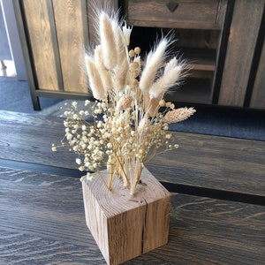 May include: A decorative floral arrangement of dried flowers in a natural wood block vase. The arrangement includes fluffy, light-coloured bunny tails, small white flowers, and wheat-like stems. The wood block has a square shape and a natural, light brown colour.