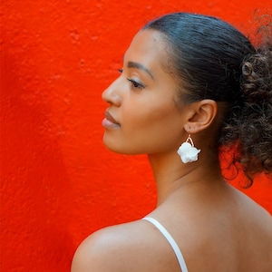 Puede incluir: Una mujer con el pelo rizado lleva un top blanco sin mangas con un lazo en la espalda. Lleva pendientes de flores blancas.