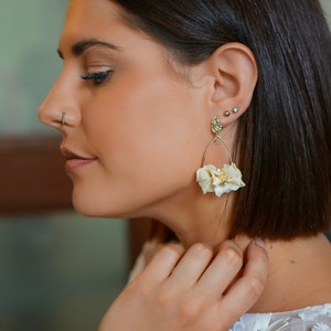 Puede incluir: Una mujer con un vestido blanco floral y un par de pendientes de gota de flores doradas. Los pendientes tienen un diseño delicado con pequeñas flores blancas.