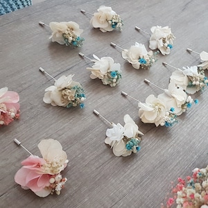 Op de afbeelding: Een verzameling van tien witte en roze gedroogde bloemstukken met blauwe accenten, elk bevestigd aan een zilveren pin. De arrangementen worden weergegeven op een lichtgrijze ondergrond.