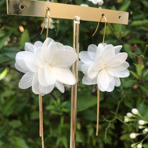 May include: A pair of white flower earrings with gold chains hanging from the bottom. The earrings are on a gold stand against a green background.