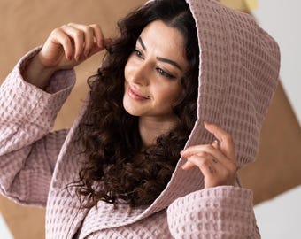 May include: A light pink waffle-weave robe with a hood and tie closure. The robe has a textured appearance and a relaxed fit. The model is wearing the robe and smiling. The robe has pockets and long sleeves.
