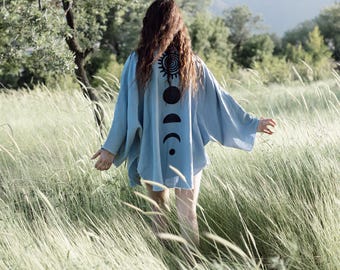 May include: A light blue kimono-style top with a black sun and moon phase design on the back. The top has wide sleeves and is worn in a field of tall grass. The image is taken outdoors on a sunny day.