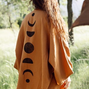 Può includere: Kimono avvolgente color senape con un design a fasi lunari nero sul retro. Il tessuto leggero e fluido è visibile nello scatto all'aperto. Il capo ha maniche ampie e una vestibilità rilassata.