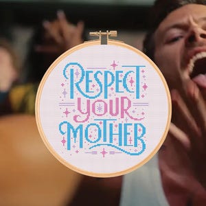 May include: A circular wooden embroidery hoop containing white fabric with cross-stitched text that reads Respect Your Mother in blue and pink lettering, accented with small pink and blue star-shaped symbols throughout the design.