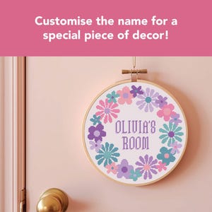 May include: A circular cross-stitch embroidery hoop featuring a floral design in pink, purple, and teal. The text "OLIVIA'S ROOM" is stitched in purple. The text above says "Customise the name for a special piece of decor!"